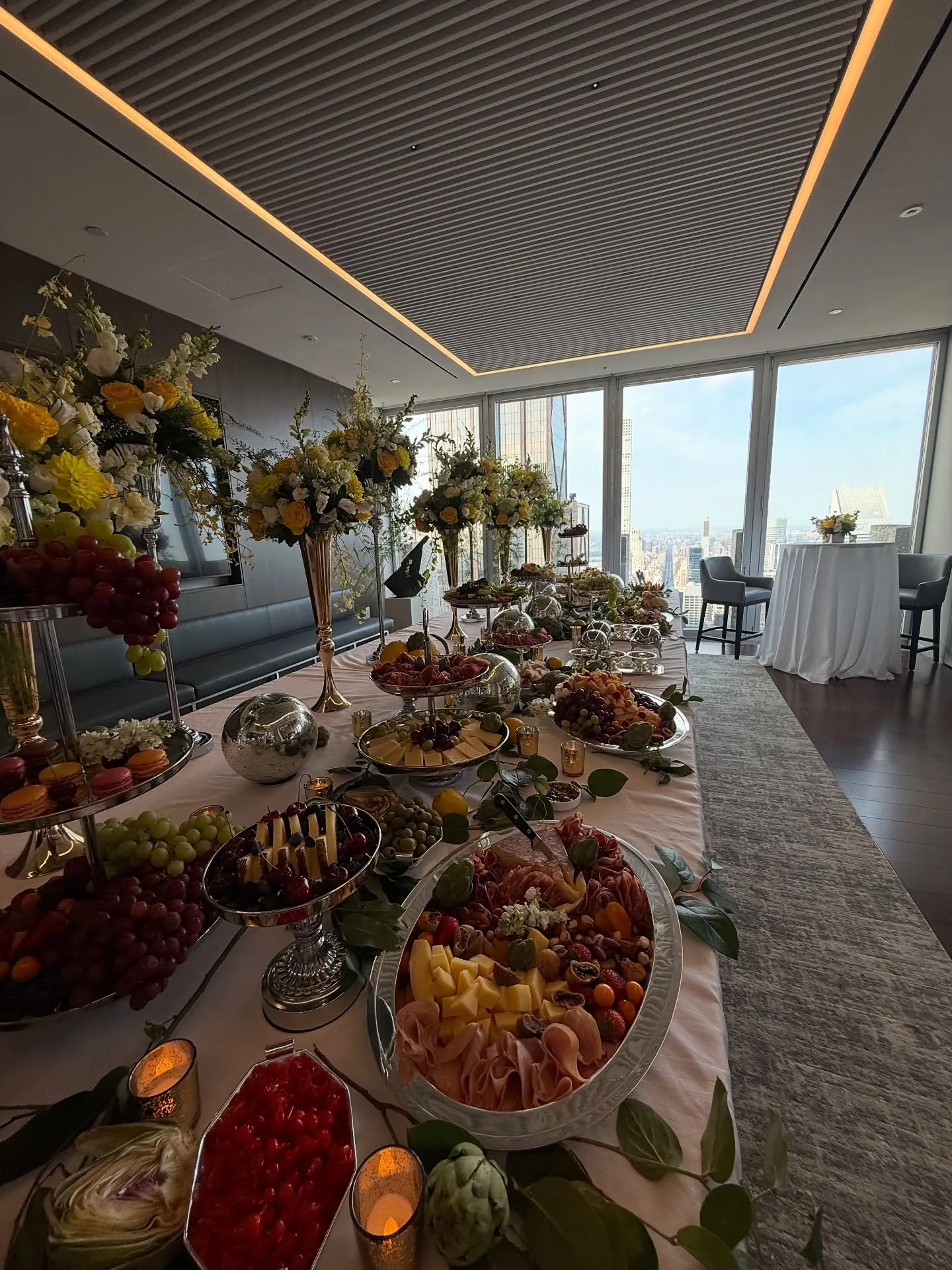 A long buffet table with charcuterie, cheese and fruit, tall yellow and white flowers and candles, in a room high above Manhattan